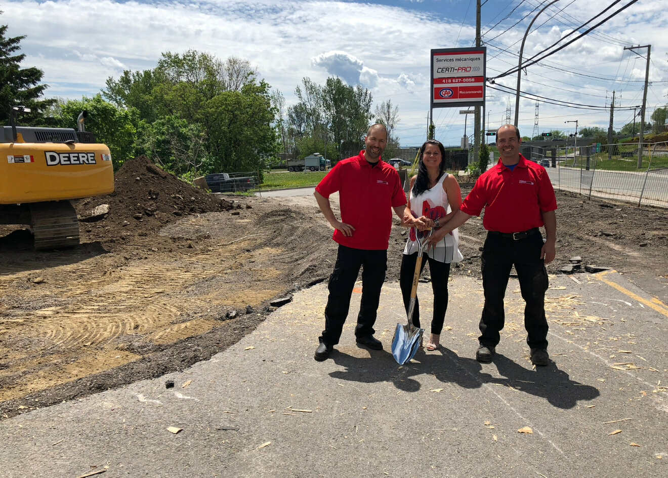 Trois personnes en vêtements rouges posent sur un chantier, tenant une pelle, avec une pelleteuse et un panneau de services en arrière-plan.