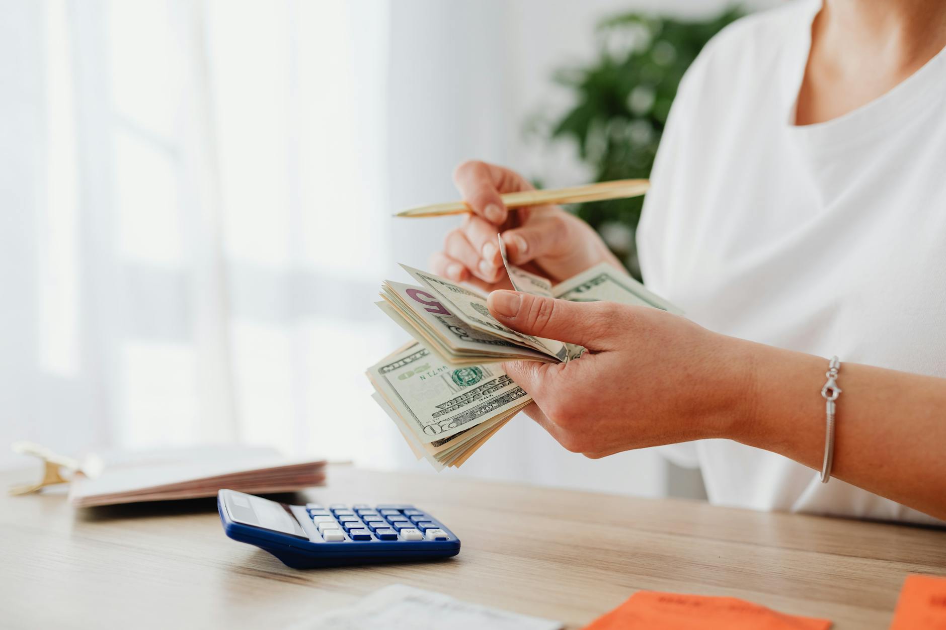 Une personne tient des billets de banque dans une main et un stylo dans l'autre, avec une calculatrice sur la table.