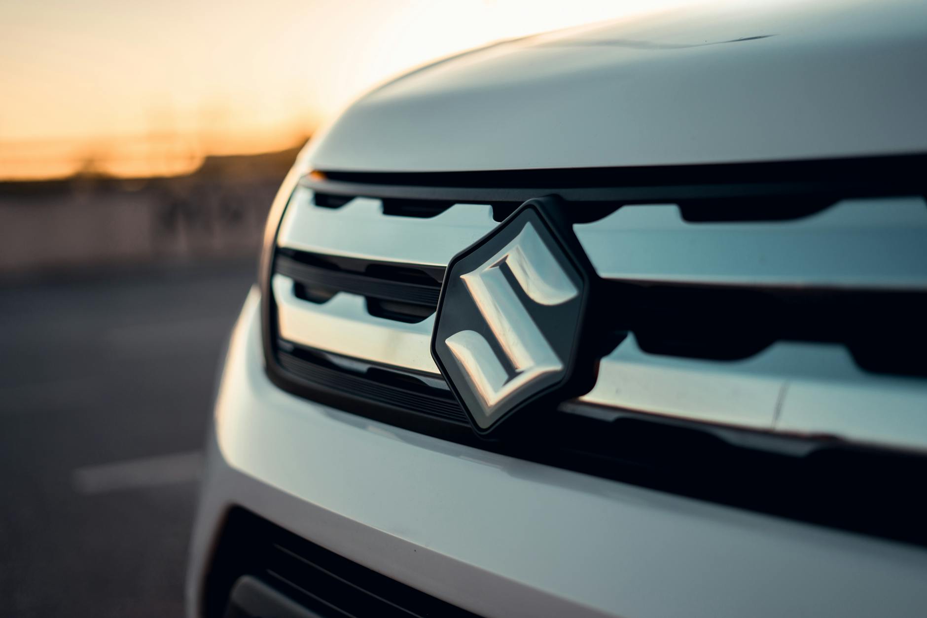 Grille avant d'un véhicule Suzuki avec logo, en arrière-plan un coucher de soleil flou.