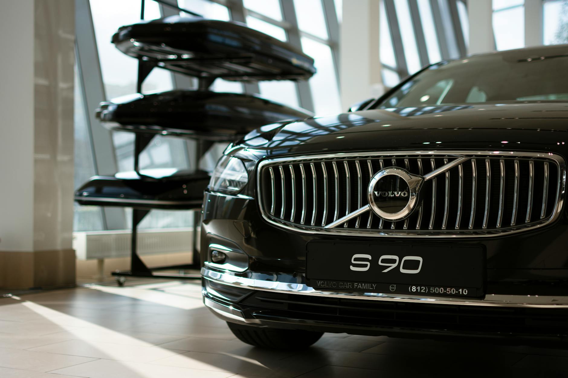 Véhicule Volvo S90 noir stationné à l'intérieur, avec des pièces de rechange exposées en arrière-plan. Éclairage naturel à travers de grandes fenêtres.
