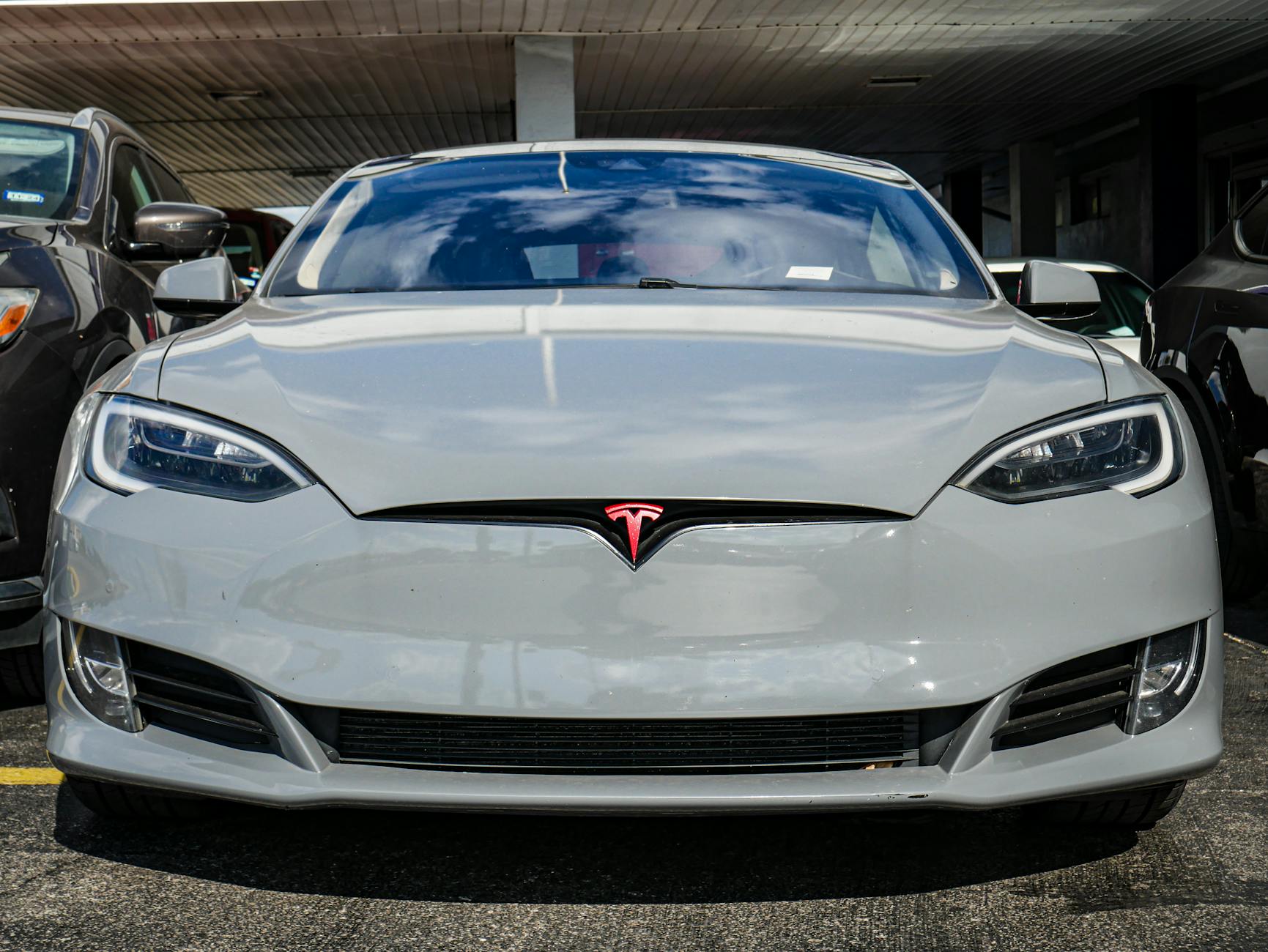 Véhicule Tesla gris stationné, vue de face, avec logo rouge sur la calandre et un environnement de stationnement en arrière-plan.