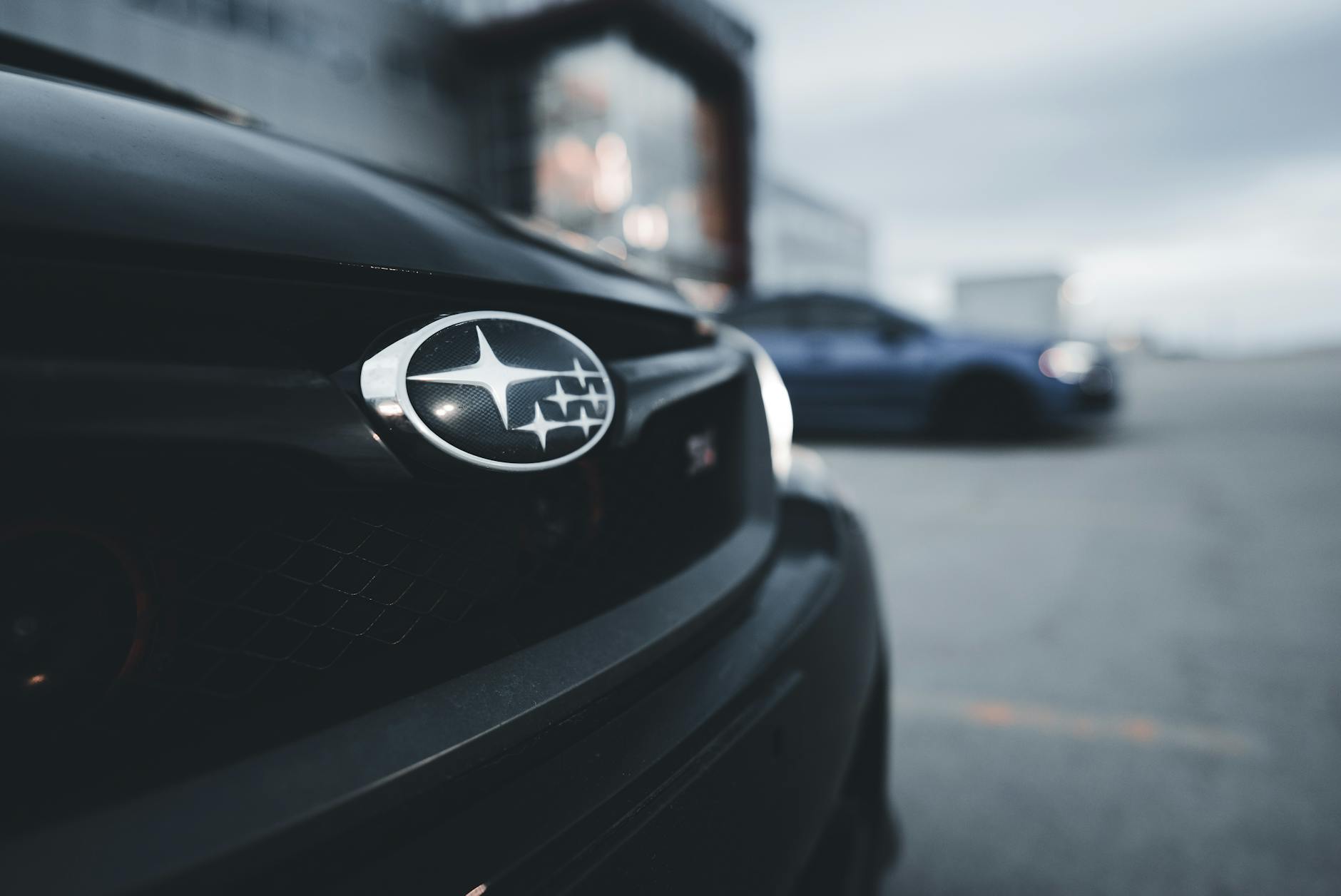 Logo Subaru en gros plan sur la calandre d'une voiture noire, avec un véhicule bleu flou en arrière-plan. Ambiance extérieure urbaine.