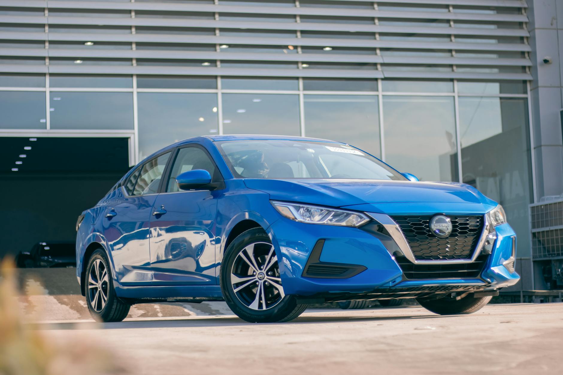 Véhicule Nissan bleu stationné devant un bâtiment moderne avec des fenêtres, sur une surface de stationnement.