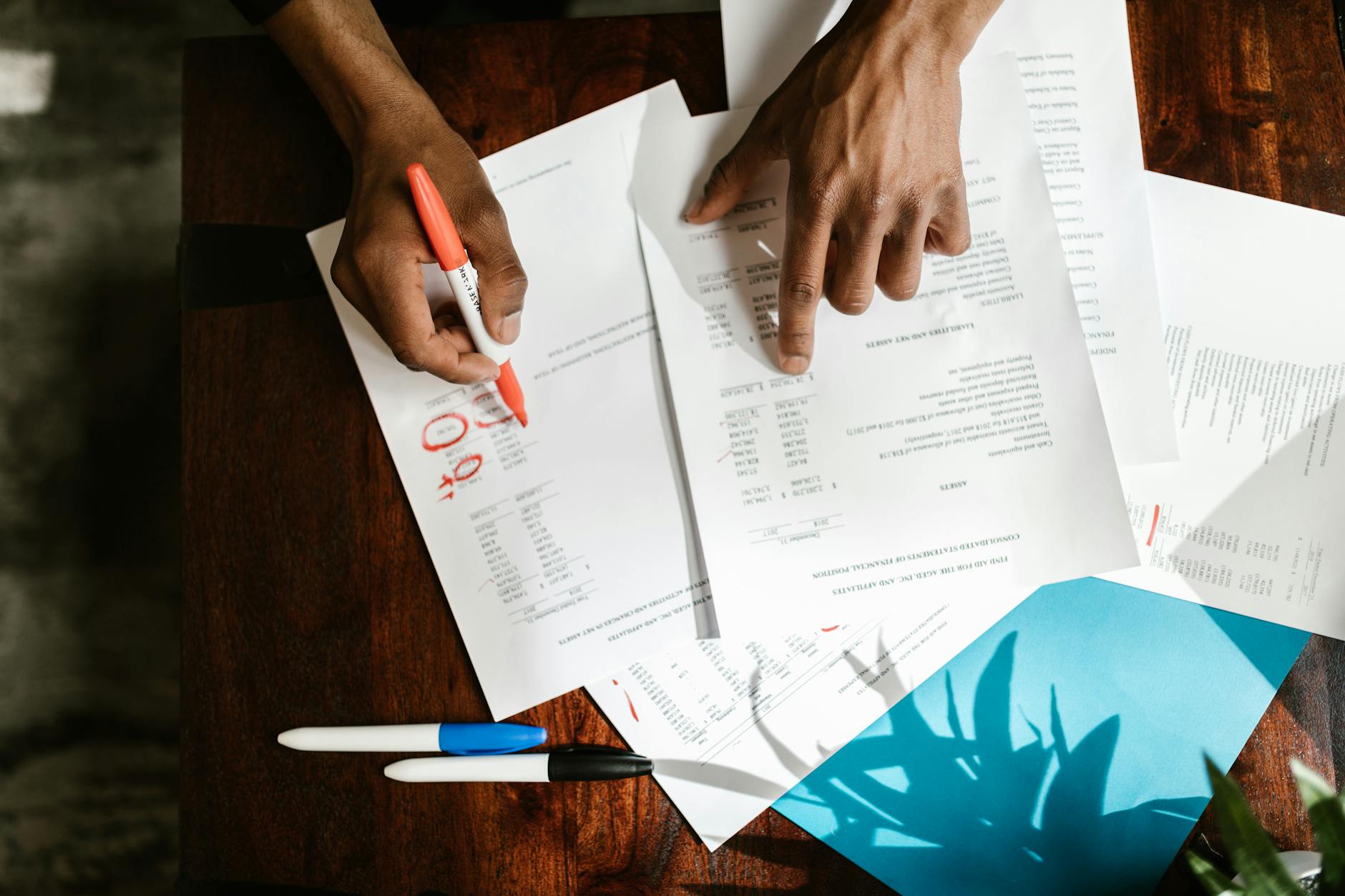 Deux mains annotent des documents financiers sur une table en bois, entourés de stylos et d'une feuille de papier colorée.