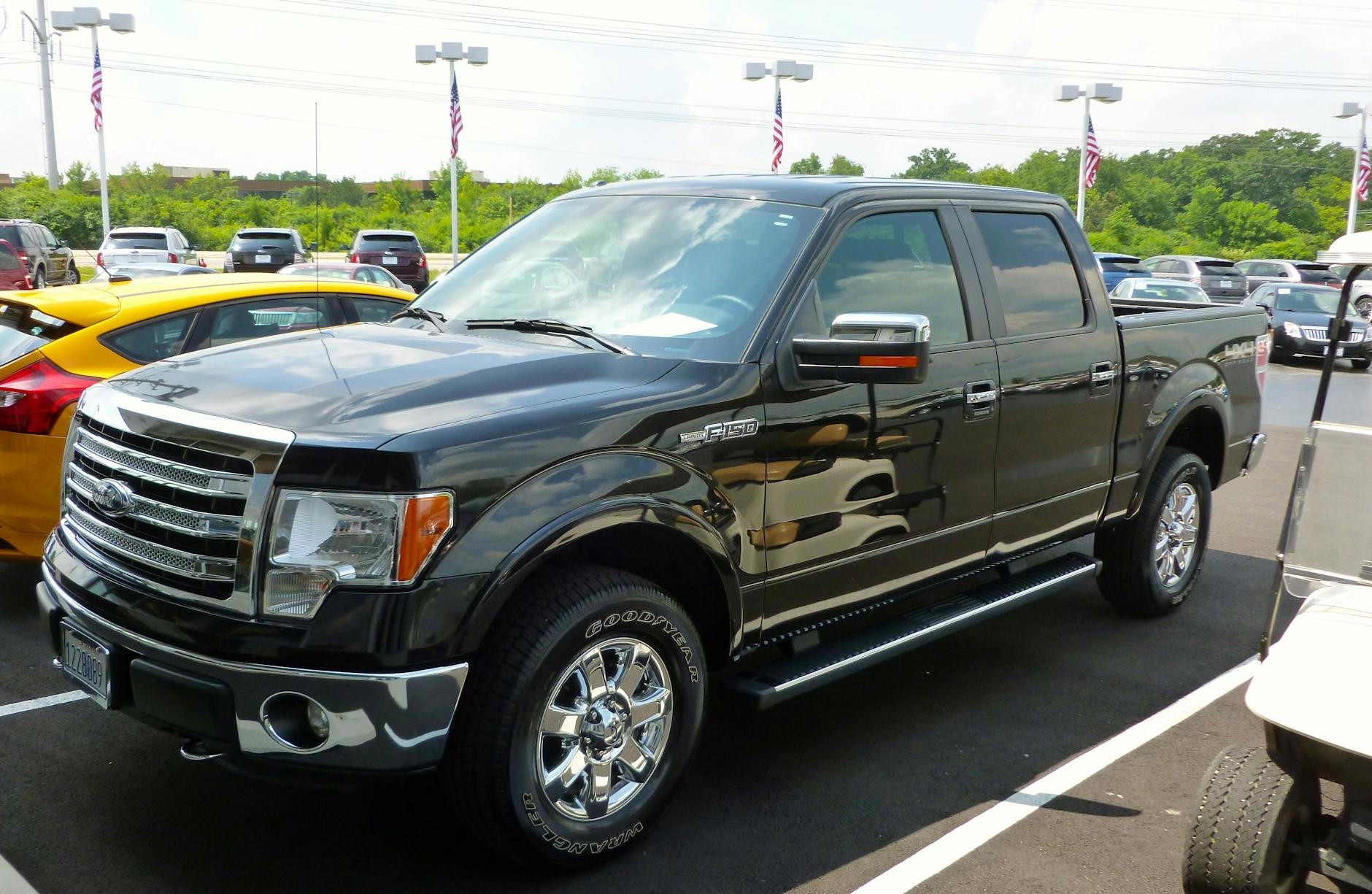 Un camion Ford F-150 noir stationné sur un terrain de vente automobile, entouré d'autres véhicules et de drapeaux américains.