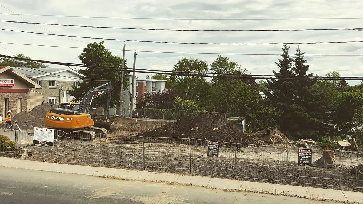 Un terrain de construction avec une excavatrice, des tas de terre et des panneaux de signalisation entourés de clôtures. Ciel nuageux en arrière-plan.
