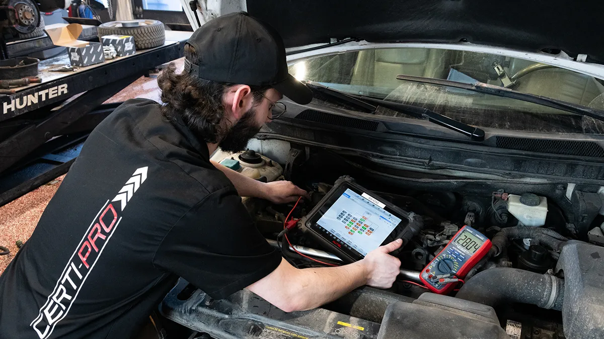 Un technicien inspecte le moteur d'un véhicule à l'aide d'une tablette et d'un multimètre, dans un garage.