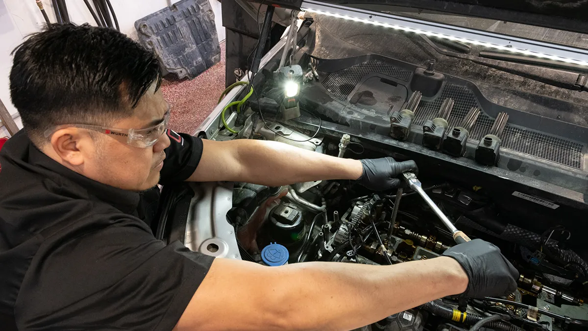 Un technicien en mécanique travaille sur un moteur de véhicule, utilisant une clé à molette et portant des gants et des lunettes de protection.