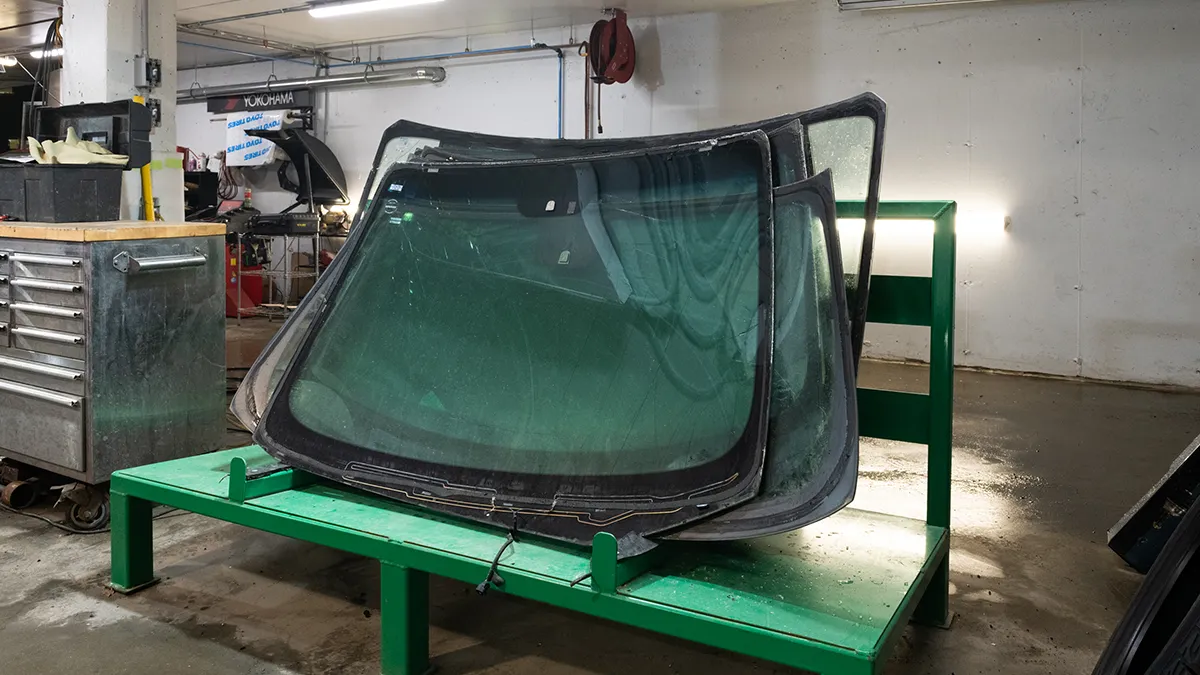 Un pare-brise de voiture posé sur un support vert, entouré d'un atelier avec des outils et équipements visibles en arrière-plan.