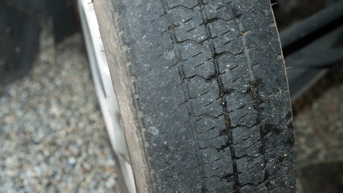 Pneu usé avec des rainures peu profondes et des signes de détérioration, posé sur un sol gravillonné.