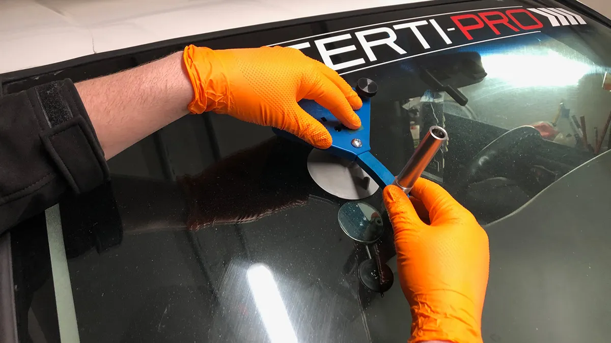 Un bras ganté orange utilise un outil bleu pour effectuer une réparation sur un pare-brise, avec un logo visible en haut de l'image.