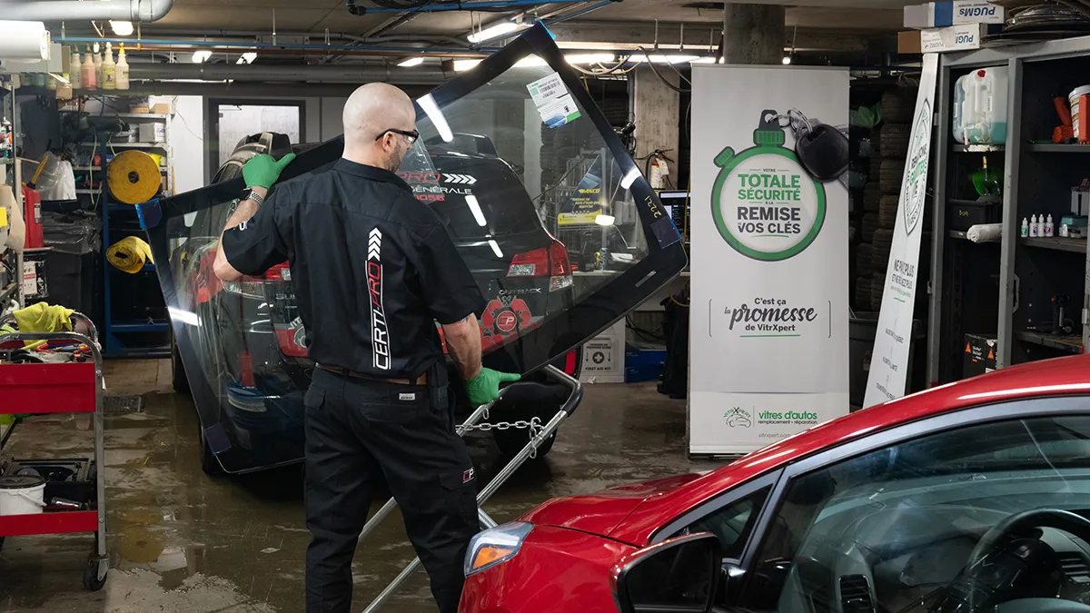 Un technicien en uniforme manipule une vitre dans un garage, avec des outils et un véhicule rouge en arrière-plan.