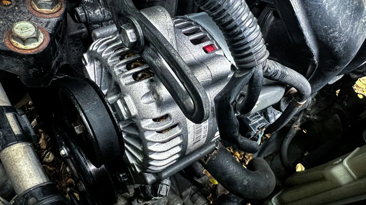 Alternateur métallique installé dans un compartiment moteur, avec des câbles et des tuyaux visibles autour.