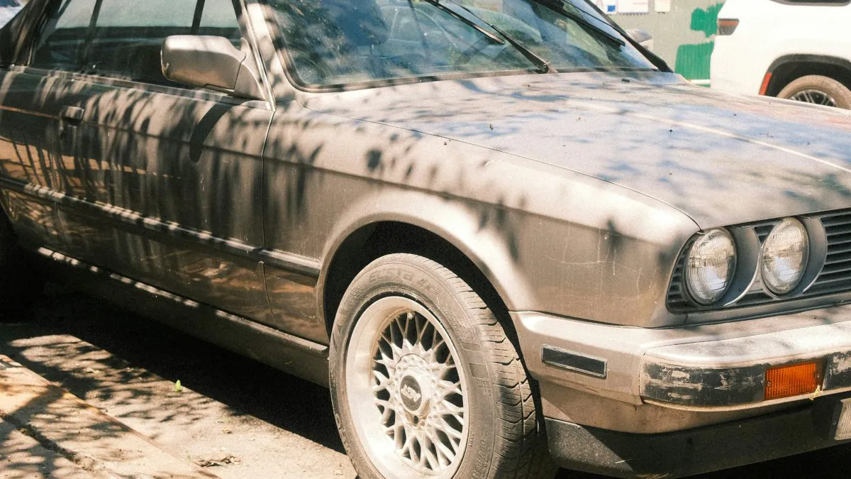 Véhicule usagé sale stationné sur une rue, avec des ombres d'arbres projetées sur sa carrosserie. Les pneus sont usés.