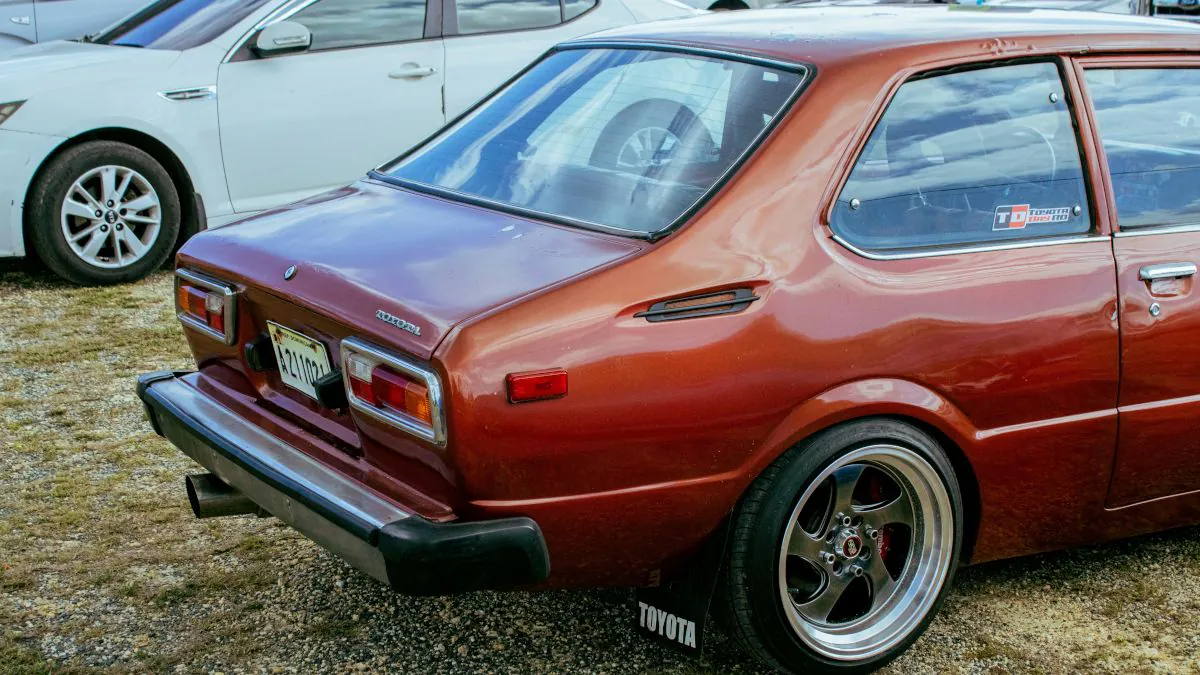 Voiture usagée de couleur rouge, vue de l'arrière, avec des jantes sport et un autocollant sur la vitre arrière. Terrain de stationnement en arrière-plan.